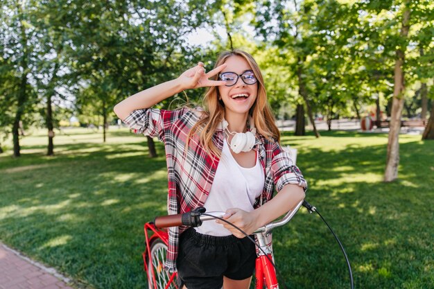 Chica positiva de moda que expresa felicidad en el parque de verano. Retrato al aire libre de dama dichosa en camisa roja posando con bicicleta.
