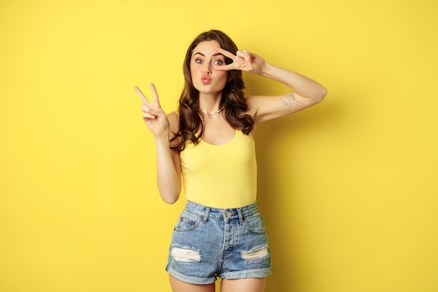 Chica positiva, modelo femenino que muestra paz, gesto de signo v y sonriente, de pie en la parte superior del tanque y pantalones cortos de mezclilla, fondo amarillo