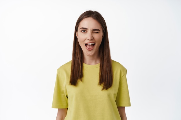 Una chica positiva te guiña un ojo, insinuando algo, sonriendo y luciendo feliz, de pie con una camiseta amarilla sobre fondo blanco