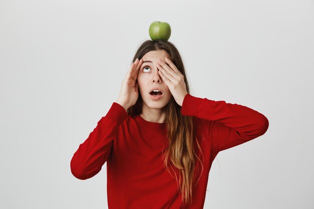 Chica preocupada asustada abre los ojos y mira el objetivo de manzana en la cabeza