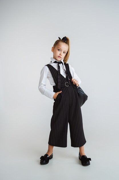 Chica preparándose para la escuela después de un largo receso de verano. De vuelta a la escuela. Pequeño modelo caucásico femenino posando en uniforme escolar con mochila sobre fondo blanco. Infancia, educación, concepto de vacaciones.