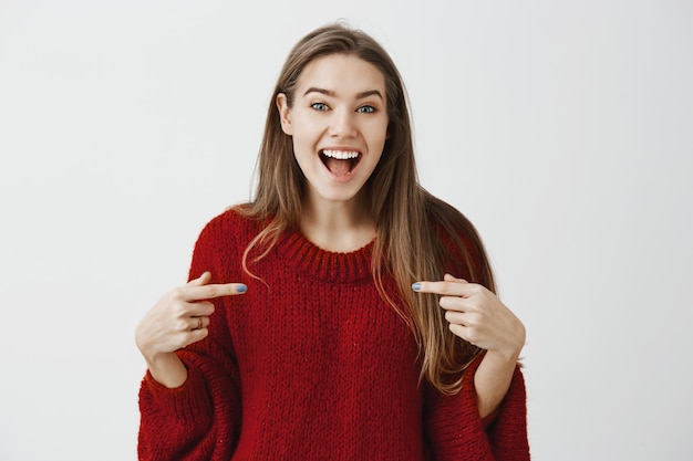 Chica presumiendo de su éxito. Foto de emocionada mujer empresaria feliz en suéter holgado de moda, apuntando con los dedos índice al pecho y sonriendo ampliamente mientras cuenta lo positivo que sucedió