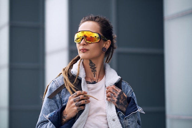 Foto gratuita chica punk con estilo con rastas, tatuajes en las manos y grandes gafas tonificadas está de pie sobre un rascacielos.