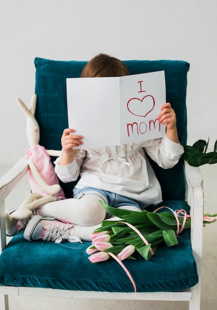 Chica que cubre la cara con la tarjeta de felicitación con la inscripción de I love mom