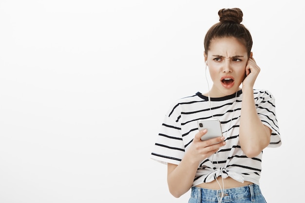 Chica que se queja disgustada que parece decepcionada, escucha música horrible o podcast en auriculares, sostiene el teléfono móvil