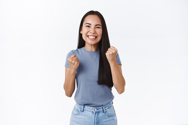 Chica que se siente feliz, aliviada a medida que se muestra la oportunidad de lograrlo. Atractiva mujer asiática sonriente bomba de puño, triunfando diciendo que sí, de pie pared blanca satisfecha con un gran resultado