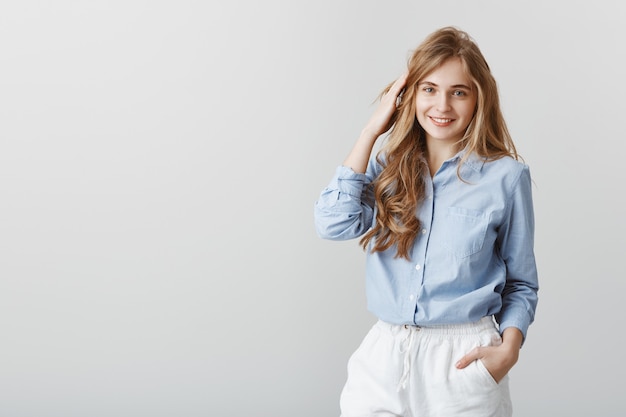 Chica que tiene una charla ordinaria con amigos. Encantadora joven rubia caucásica en blusa azul, tocando el cabello y sonriendo alegremente, siendo educado con el cliente durante el trabajo en la oficina