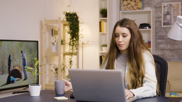 Chica que trabaja en la computadora portátil desde la oficina en casa. Chica tomando un sorbo de café.