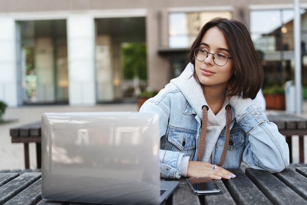 Chica queer romántica de ensueño con corte de pelo corto, mirando a la izquierda con una sonrisa encantadora y pensativa, usando un teléfono móvil y una computadora portátil, trabajando al aire libre, estudiando en el parque, esperando un amigo, concepto de estilo de vida.