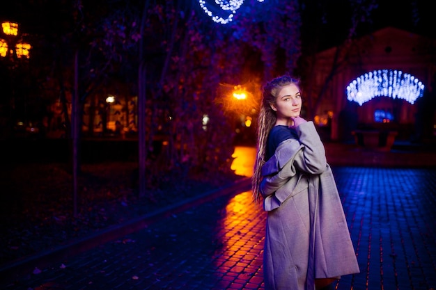 Chica con rastas caminando por la calle nocturna de la ciudad contra luces de guirnaldas