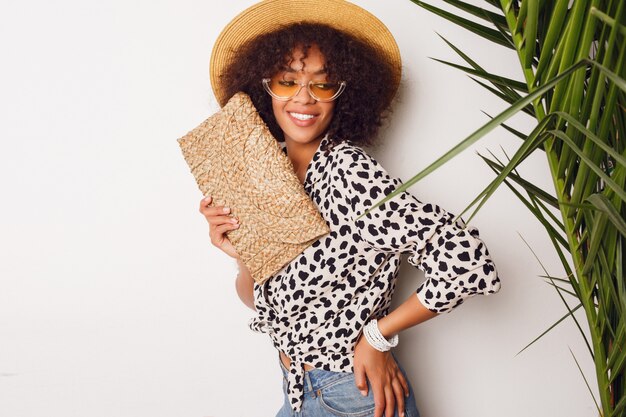 Chica de raza mix de moda en blusa de moda y sombrero de paja de pie sobre la pared blanca. Perfecta sonrisa blanca. Humor de compras