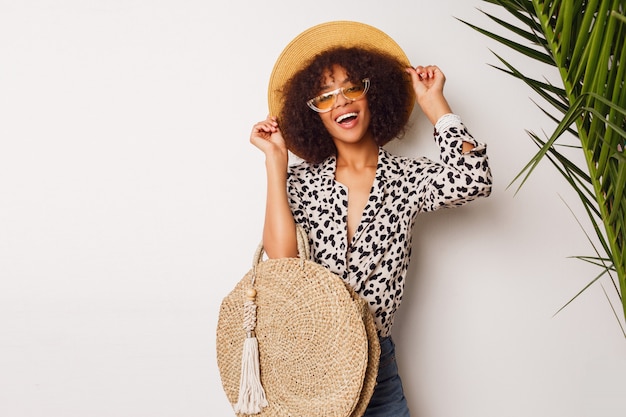 Chica de raza mix de moda en blusa de moda y sombrero de paja de pie sobre la pared blanca. Perfecta sonrisa blanca. Humor de compras