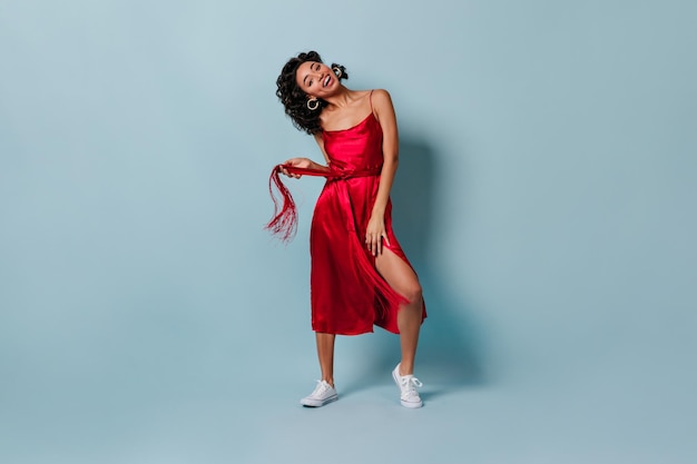 Chica de raza mixta alegre en vestido rojo mirando a la cámara Vista de longitud completa de mujer joven sensual con cabello rizado posando sobre fondo azul