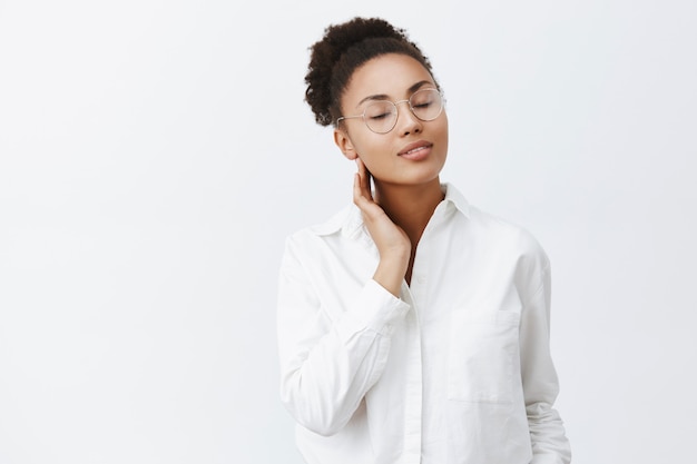 Chica recuerda apasionado beso de amante. Retrato de mujer afroamericana femenina tierna con gafas y camisa blanca, cerrando los ojos y tocando el cuello suavemente, de pie sobre la pared gris