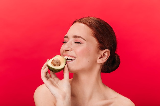 Chica refinada comiendo aguacate Foto de estudio de encantadora mujer caucásica posando sobre fondo rojo