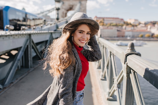 Chica refinada en elegante abrigo de tweed posando con una sonrisa encantadora sobre fondo urbano durante el viaje