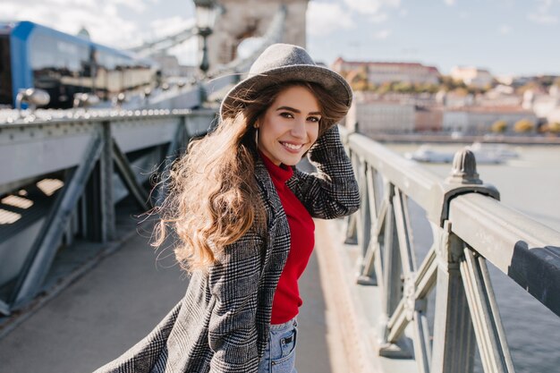 Chica refinada en elegante abrigo de tweed posando con una sonrisa encantadora sobre fondo urbano durante el viaje