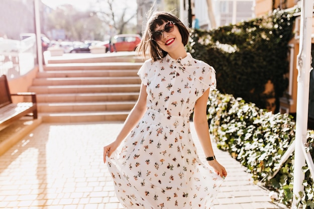 Chica refinada con labios rojos sonriendo durante la caminata en día de verano. Mujer de pelo castaño con vestido blanco largo que expresa felicidad en la ciudad.