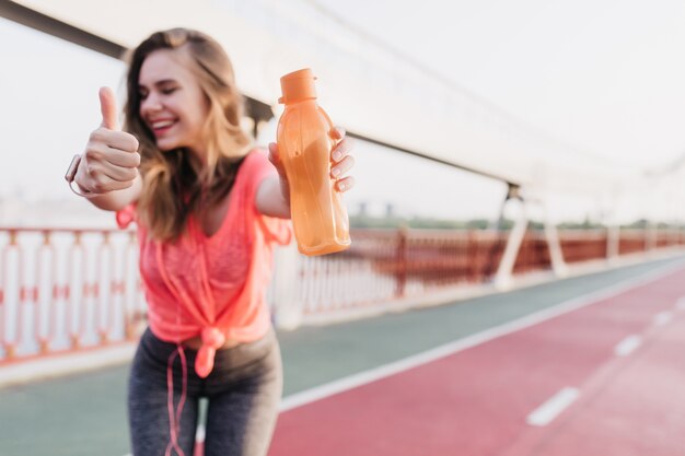 Chica refinada en pantalones deportivos grises posando con una sonrisa. Risa agraciada mujer de pie con botella en el estadio.