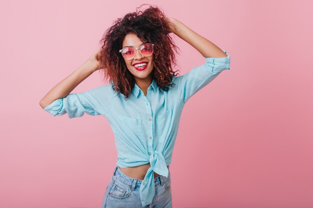 Chica refinada con piel negra disfrutando de la moda. Preciosa dama mulata jugando con el pelo rizado y expresando felicidad.