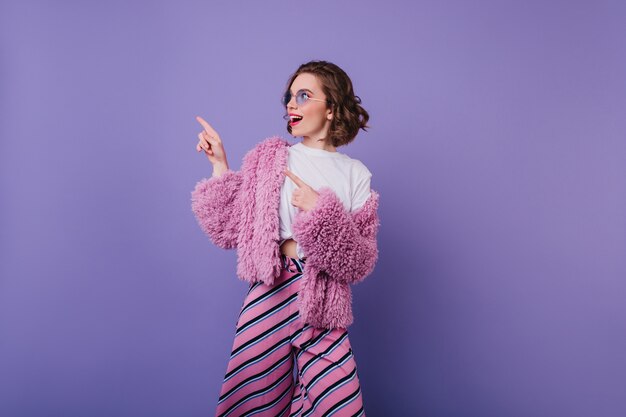 Chica relajada en pantalones de rayas rosas divirtiéndose. Modelo de mujer de ensueño lleva abrigo de piel y gafas de sol bailando en la pared púrpura.