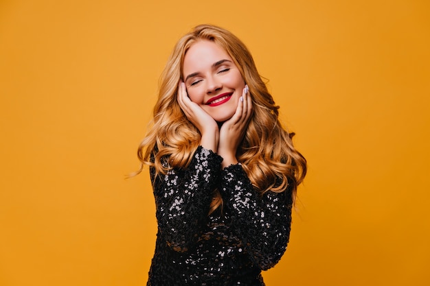 Chica relajada en vestido negro posando con una sonrisa. dama rubia despreocupada aislada en la pared amarilla.