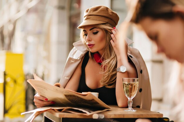 Chica relajante en ropa elegante leyendo el periódico después del trabajo, sentada en la cafetería con una copa de champán. Retrato de mujer adorable con maquillaje desnudo en reloj de pulsera escalofriante en restaurante.