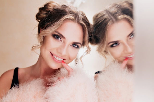 Chica riendo alegre con peinado lindo posando mientras hace maquillaje junto al espejo. Retrato de mujer joven rizada alegre en abrigo de moda de pie con una sonrisa sincera sobre fondo claro