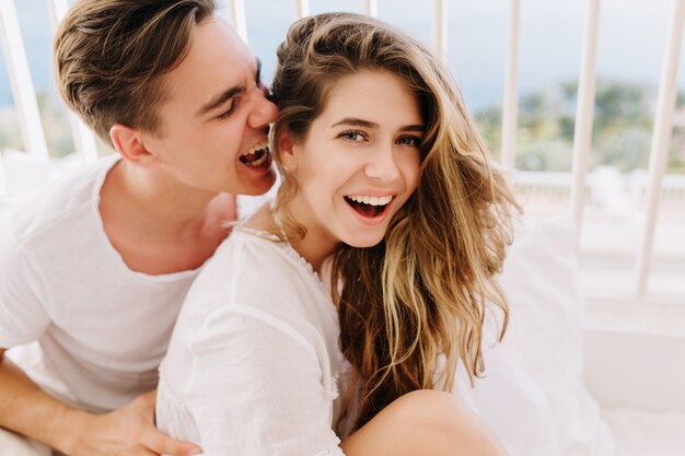 Chica riendo alegre con peinado de moda divirtiéndose con su marido en traje blanco el domingo por la mañana. Hermosa pareja de buen humor pasar tiempo juntos en vacaciones, sentados en la cama