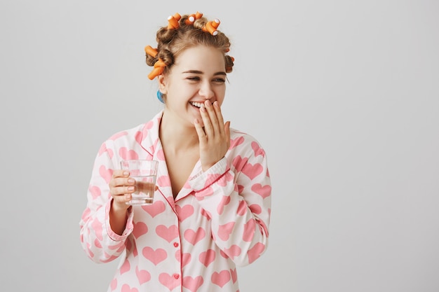 Chica riendo alegre en rizadores de pelo y pijama bebiendo un vaso de agua