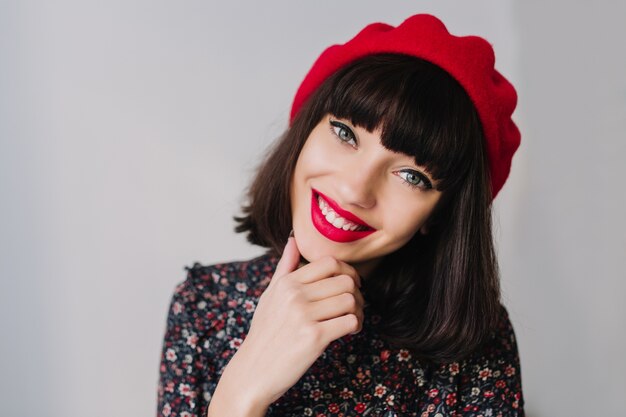 Chica riendo con peinado corto y traje francés de moda posando tocando su barbilla sobre fondo borroso. Close-up retrato de sonriente joven morena en ropa vintage mirando con admiración