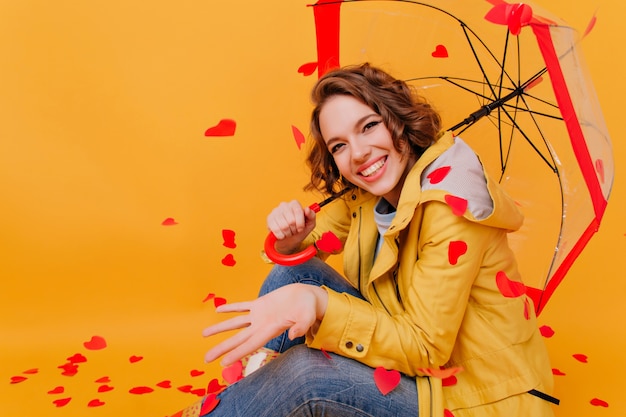 Chica riendo refinada en chaqueta amarilla de moda posando bajo la sombrilla. Retrato de estudio de mujer agraciada de buen humor sentada en el suelo con corazones en la pared.