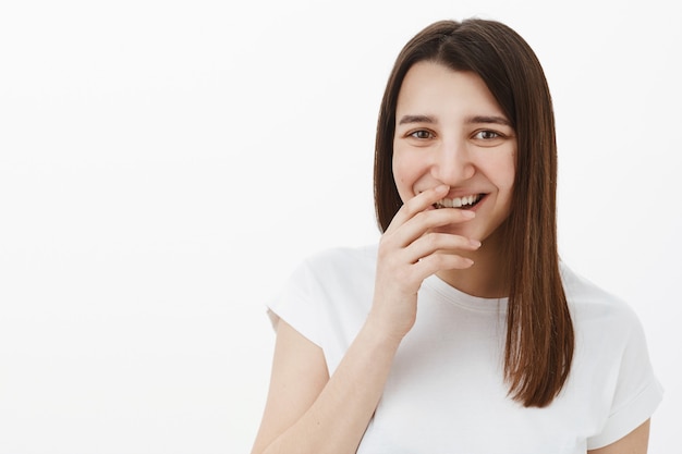 Chica riéndose de ti mientras te diviertes y bromeando estando en un estado de ánimo alegre y juguetón riendo tontamente, cubriendo una sonrisa perfecta con la mano y mirando divertido posando en camiseta blanca contra la pared gris