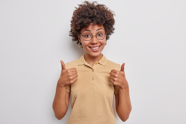 La chica rizada alegre mantiene los pulgares arriba expresa me gusta y la retroalimentación positiva aprueba y apoya el plan alaba la venta en la tienda usa una camiseta casual aislada sobre fondo blanco celebra un buen resultado