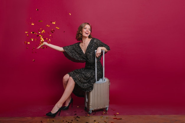 Chica rizada alegre en vestido negro retro descansando después de empacar la maleta para el próximo viaje