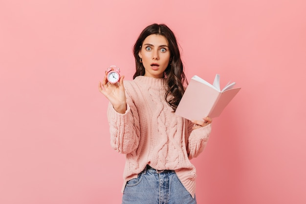 Chica rizada con asombro mira a cámara sobre fondo rosa. Mujer en suéter con libro y despertador.