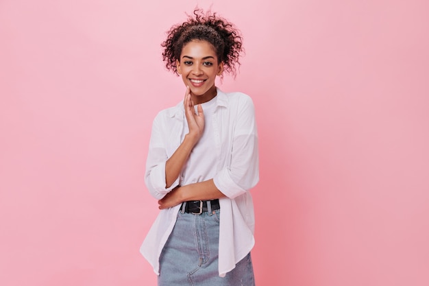 Chica rizada en camisa blanca está sonriendo en la pared rosa