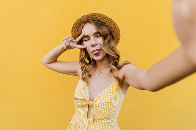 Chica rizada divertida con sombrero de paja haciendo selfie con signo de la paz. Foto interior de una magnífica dama rubia jugando.