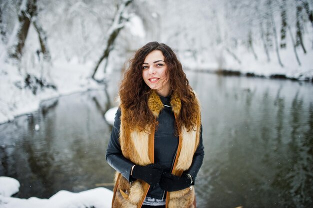 Chica rizada de elegancia en abrigo de piel en el parque forestal nevado contra el río congelado en invierno