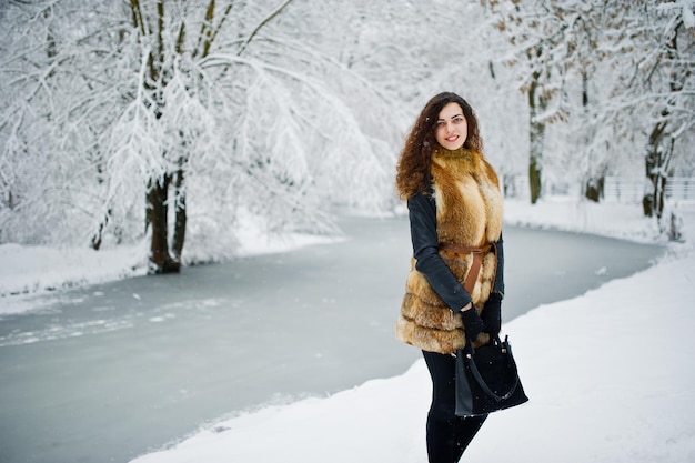 Chica rizada de elegancia en abrigo de piel en el parque forestal nevado en invierno