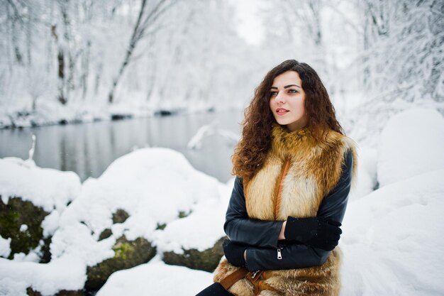 Chica rizada de elegancia en abrigo de piel en el parque forestal nevado en invierno