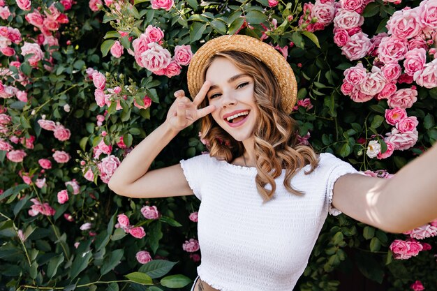 Chica rizada emocionada en traje de verano haciendo selfie. Foto al aire libre de una joven romántica divirtiéndose en el jardín.