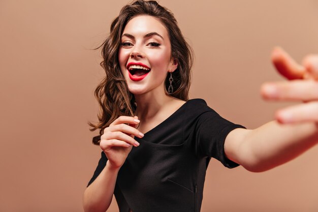 Chica rizada con labios rojos vestida con top negro hace selfie sobre fondo beige.