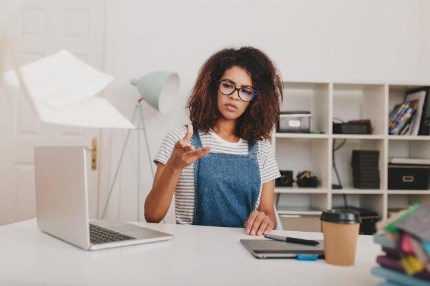 Chica rizada molesta con gafas y camisa a rayas esparce documentos sentado en la mesa y mirando a otro lado. Mujer joven infeliz que trabaja con la computadora portátil y la computadora portátil que hace su trabajo en la oficina.