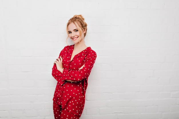 Chica rizada positiva en pijama de pie en pose de confianza. retrato de modelo femenino relajado viste camisón rojo y posando con los brazos cruzados.