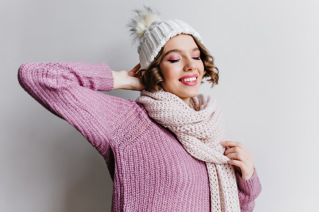 Chica rizada romántica con sombrero blanco con caja de regalo rosa y sonriendo. Señora alegre con corte de pelo corto mirando el regalo de Navidad con expresión de cara feliz.