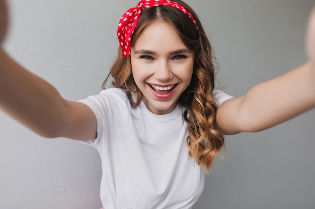 Foto gratuita chica rizada sensual con cinta roja haciendo selfie. foto interior de refinada modelo femenina con peinado ondulado riendo