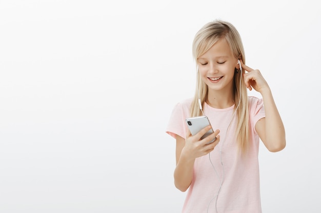 Chica robó el teléfono de mamá para ver la nueva serie de dibujos animados favoritos. Niña juguetona complacida con cabello rubio, escuchando música en auriculares y sonriendo en la pantalla del teléfono inteligente mientras juega