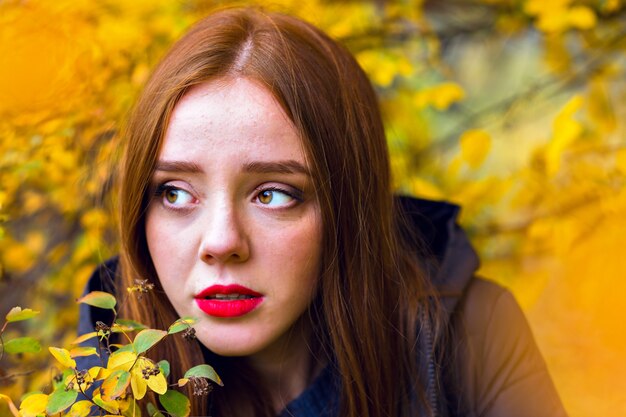 Chica romántica con cabello lacio brillante mirando a otro lado, escondiéndose detrás de un follaje amarillo. Retrato al aire libre del primer del modelo femenino morena solitario que presenta en el parque del otoño.