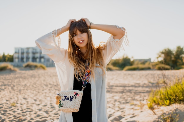 Chica romántica con cabello largo morena caminando por la playa. Estilo bohemio, bolso de paja, maquillaje brillante. Estado de ánimo tropical. Colores del atardecer.